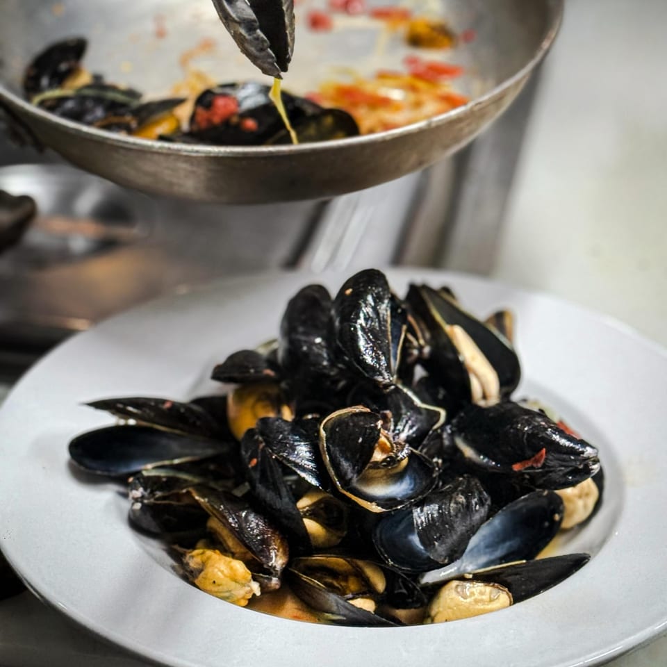 A plate of cooked mussels sits beneath a pan where tongs drizzle sauce over seafood in a commercial kitchen.