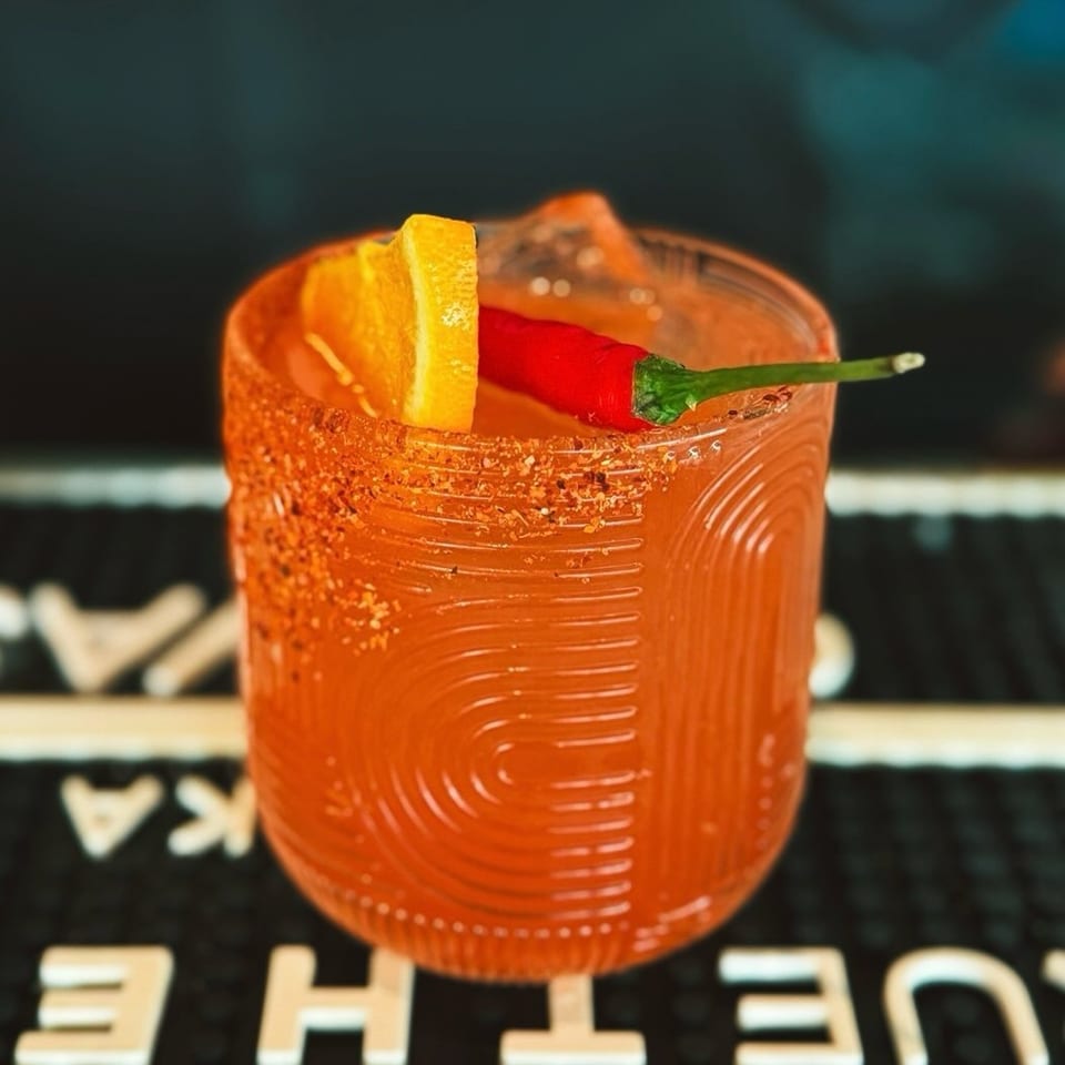 Spicy red cocktail in a textured glass with a chili pepper and orange wedge garnish on a bar counter.