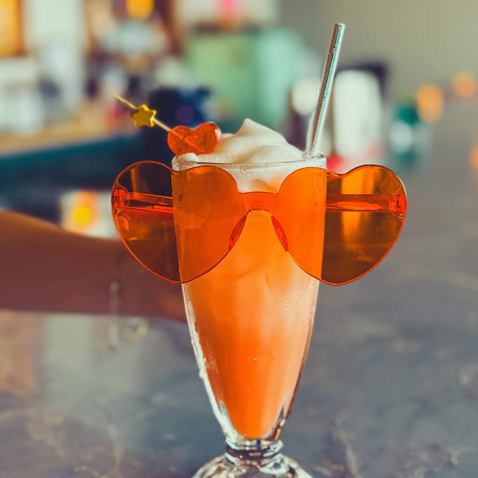 Orange cocktail topped with whipped cream, heart-shaped garnish, star pick, and straw, photographed behind oversized orange sunglasses.