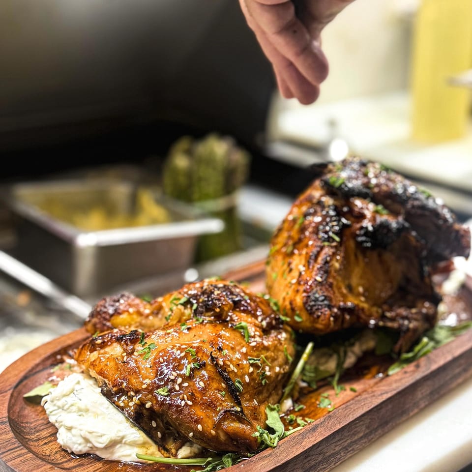 Chef garnishes glazed, sesame-crusted grilled meat with herbs on a wooden serving platter beside creamy sauce.