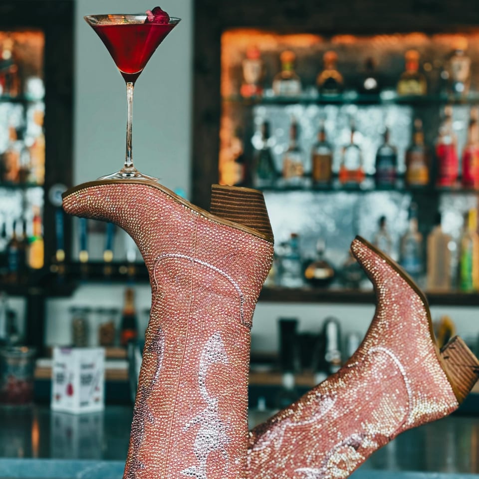 Red cocktail garnished with berries beside sparkling pink cowboy boots at the Boardwalk Restaurant bar.