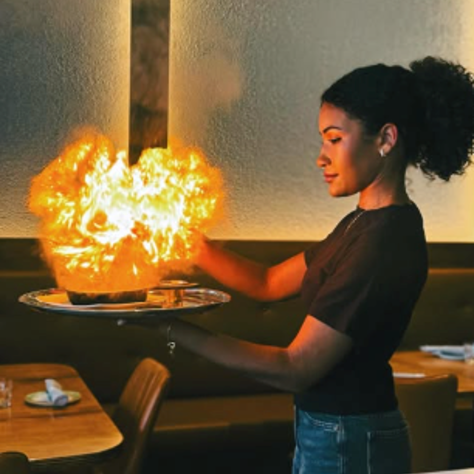 Restaurant server presents a flaming dish on a platter beside a dining table.